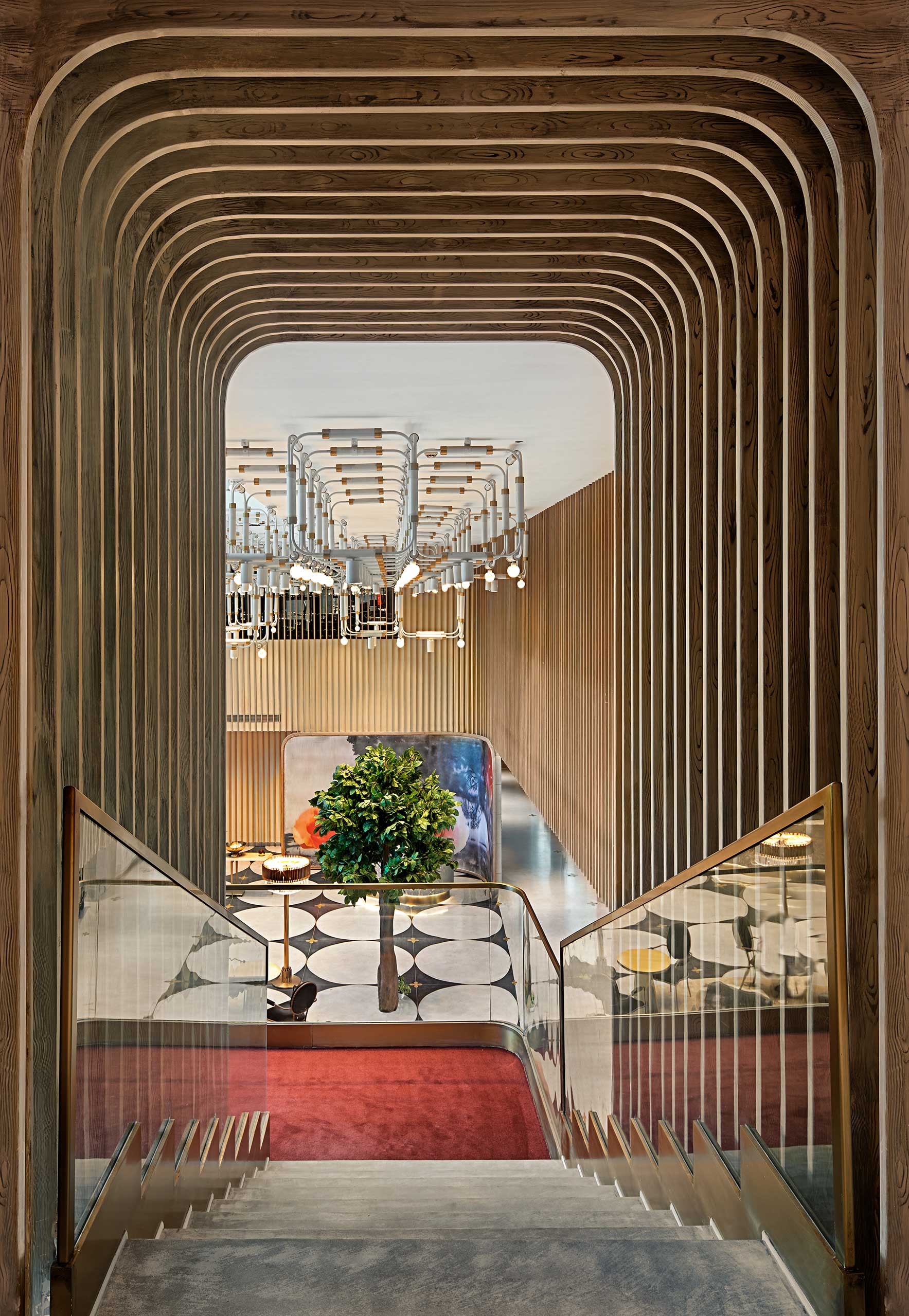 Modern hotel interior with wooden archways lining a staircase. A geometric chandelier hangs above a green tree centerpiece, set against patterned flooring.