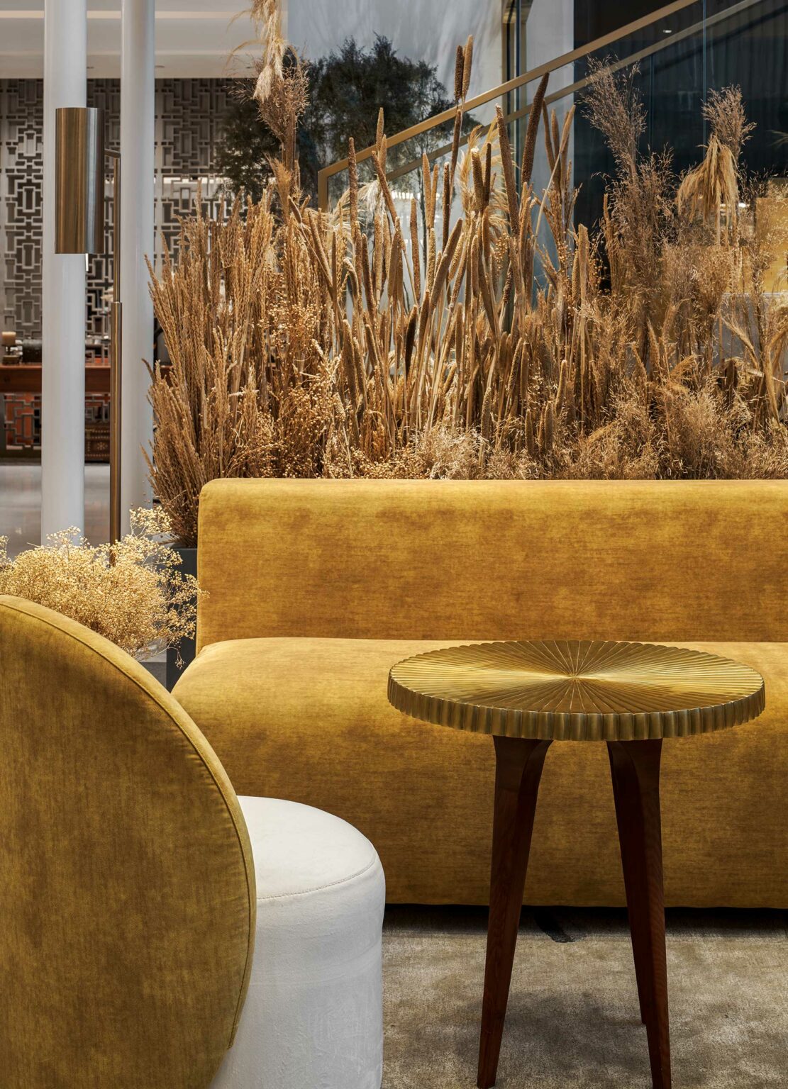 Stylish hotel interior with a mustard yellow sofa, round wooden table, and a mix of white and mustard chairs. Tall dried plants create a warm, cozy ambiance.