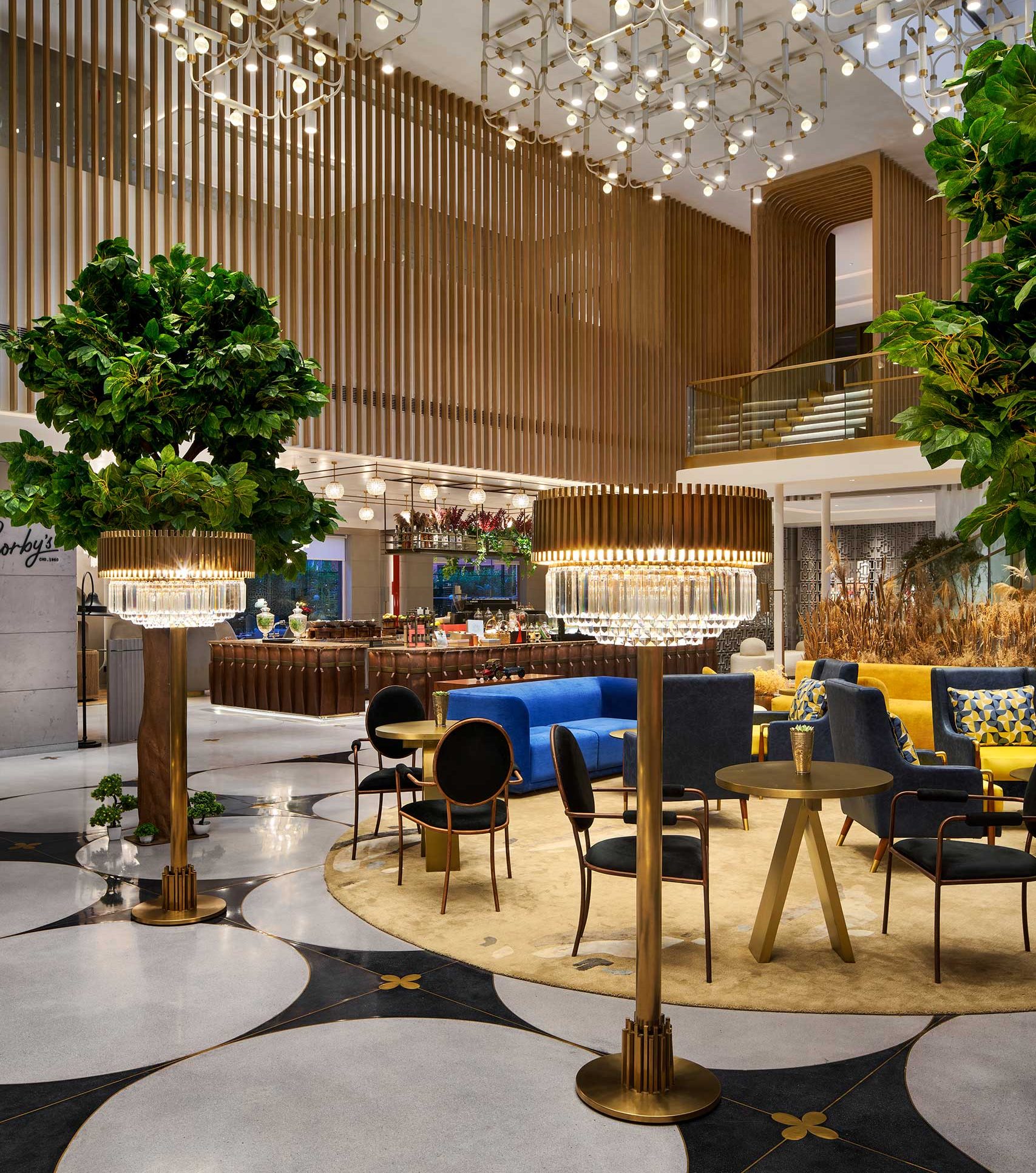 Luxurious hotel lobby with modern chandeliers, elegant seating with blue and yellow sofas, indoor trees, wooden accents, and a polished floor.