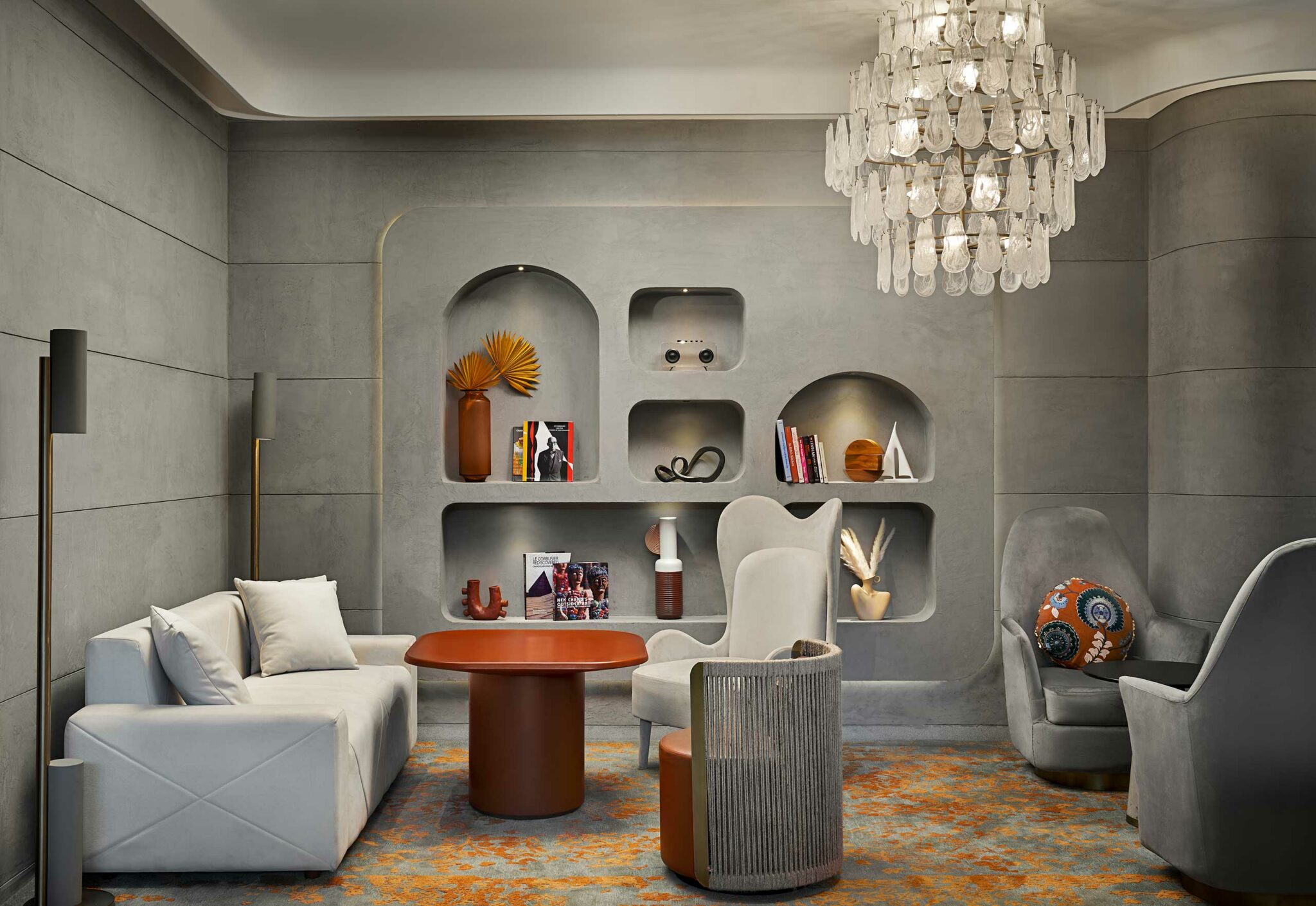 A modern, cozy hotel lounge with gray walls features a built-in shelf displaying vases and books. A chandelier hangs above plush seating and a round table.