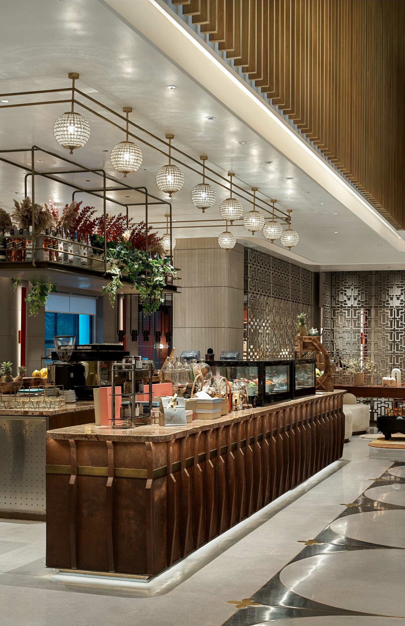 A modern hotel cafe area with a long wooden counter adorned with pastries, glass chandeliers, decorative shelving with plants and warm ambient lighting.