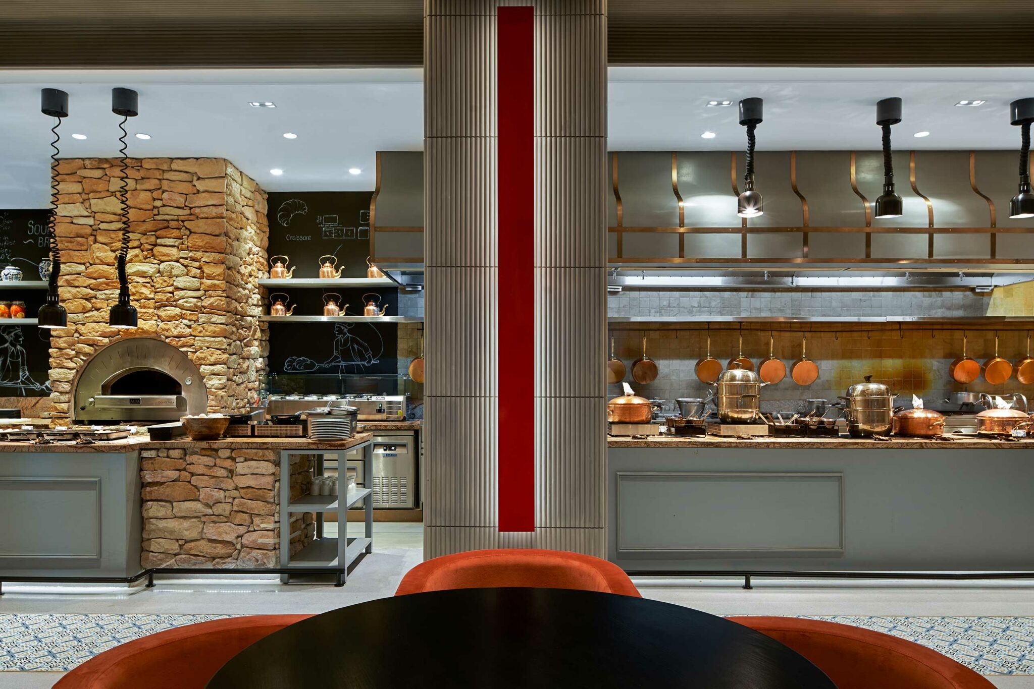 Modern hotel open kitchen area with stone pizza oven, hanging pots, and open shelving. Warm lighting creates an inviting, elegant atmosphere.
