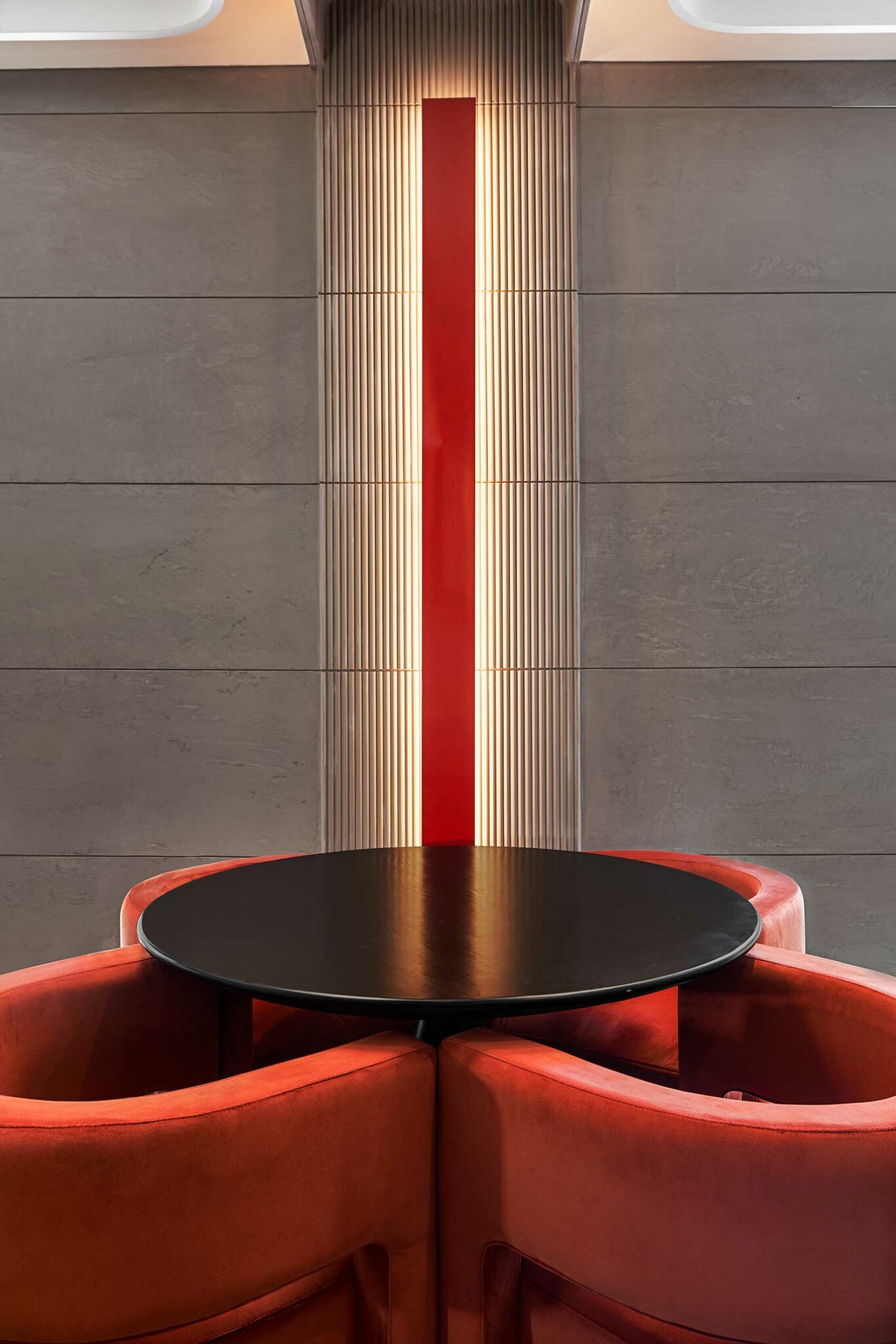 Modern hotel cafe area with a black round table surrounded by red cushioned chairs. Behind them, a vertical red light bar on a gray textured wall.