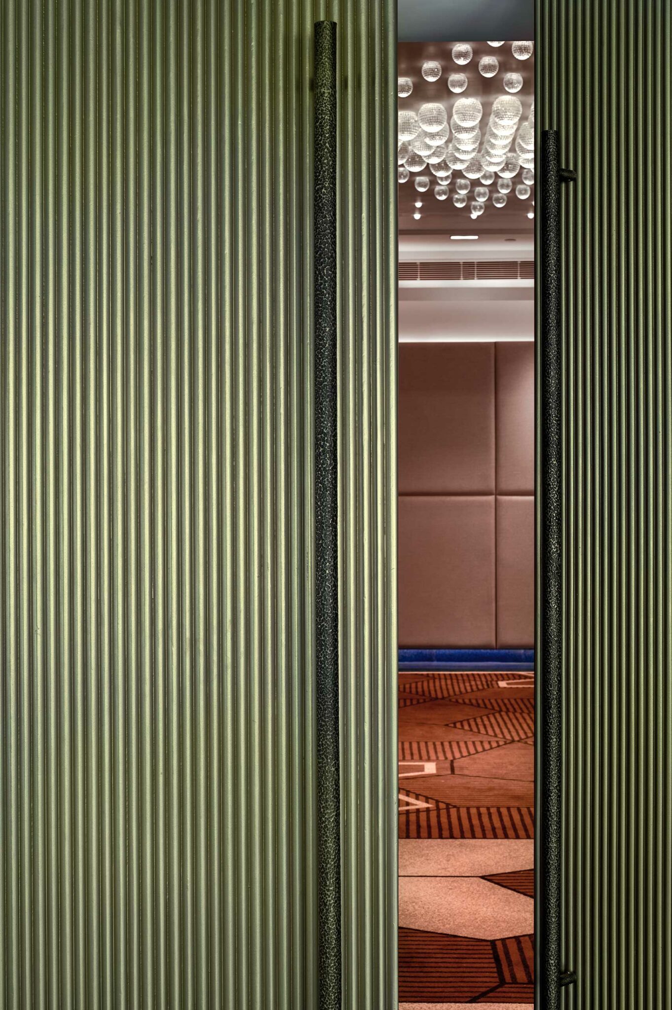 A partially open door with vertical metallic grooves reveals a sleek, modern room. Inside, a geometric carpet and unique bubble-like ceiling lights are visible.