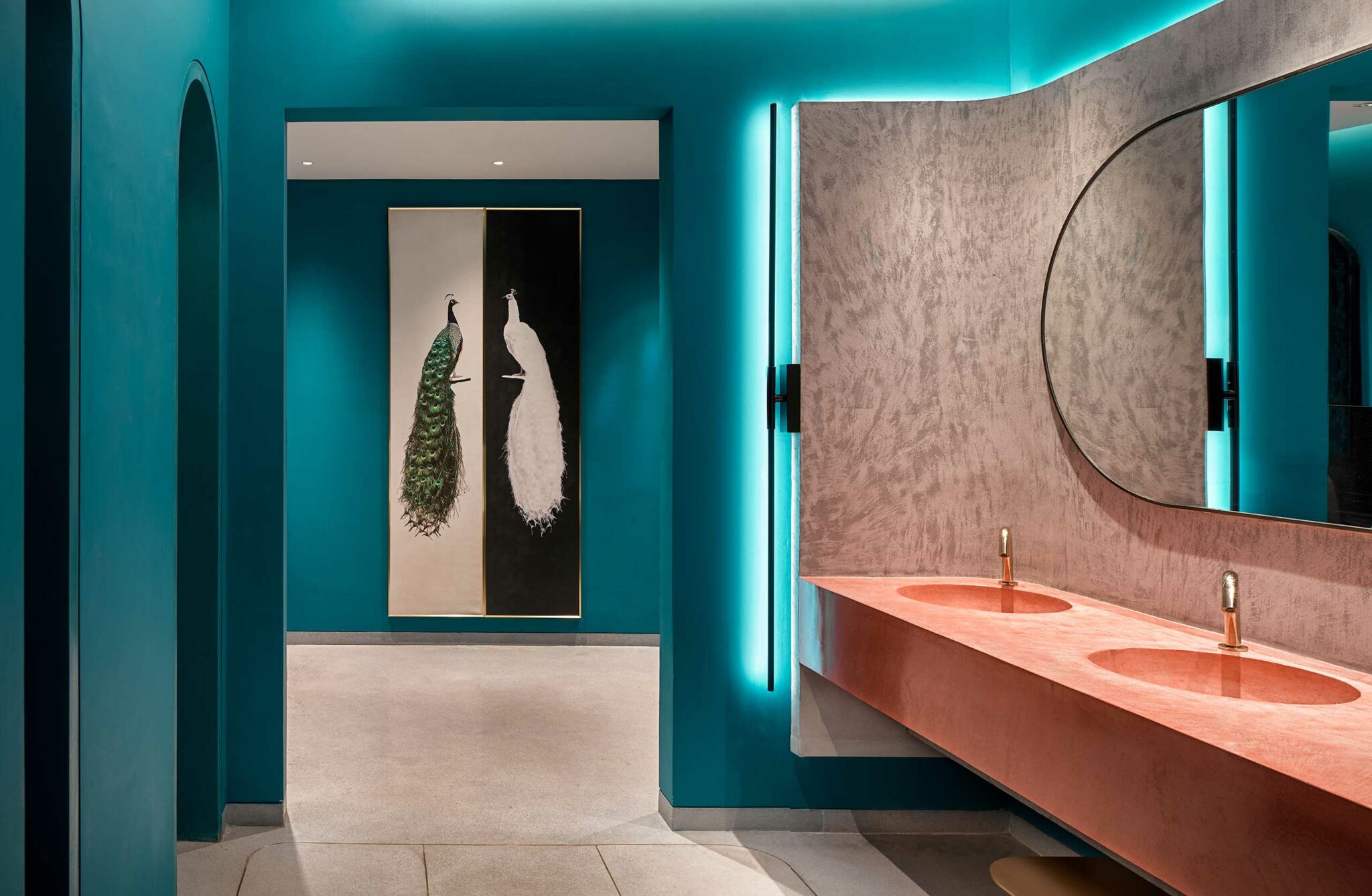 Modern hotel bathroom with teal walls, featuring a large mirror and coral sinks. A painting of two peacocks is displayed in the background, enhancing the elegant ambiance.