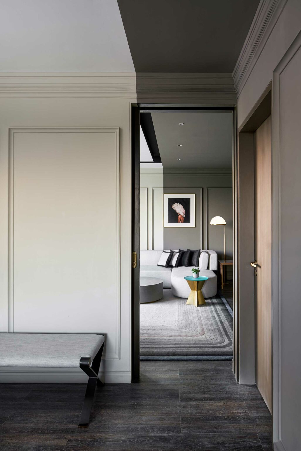 A modern hotel room with a partially open door revealing a cozy interior. It features a gray sofa, elegant floor lamp, abstract art, and a gold side table, creating a serene ambiance.