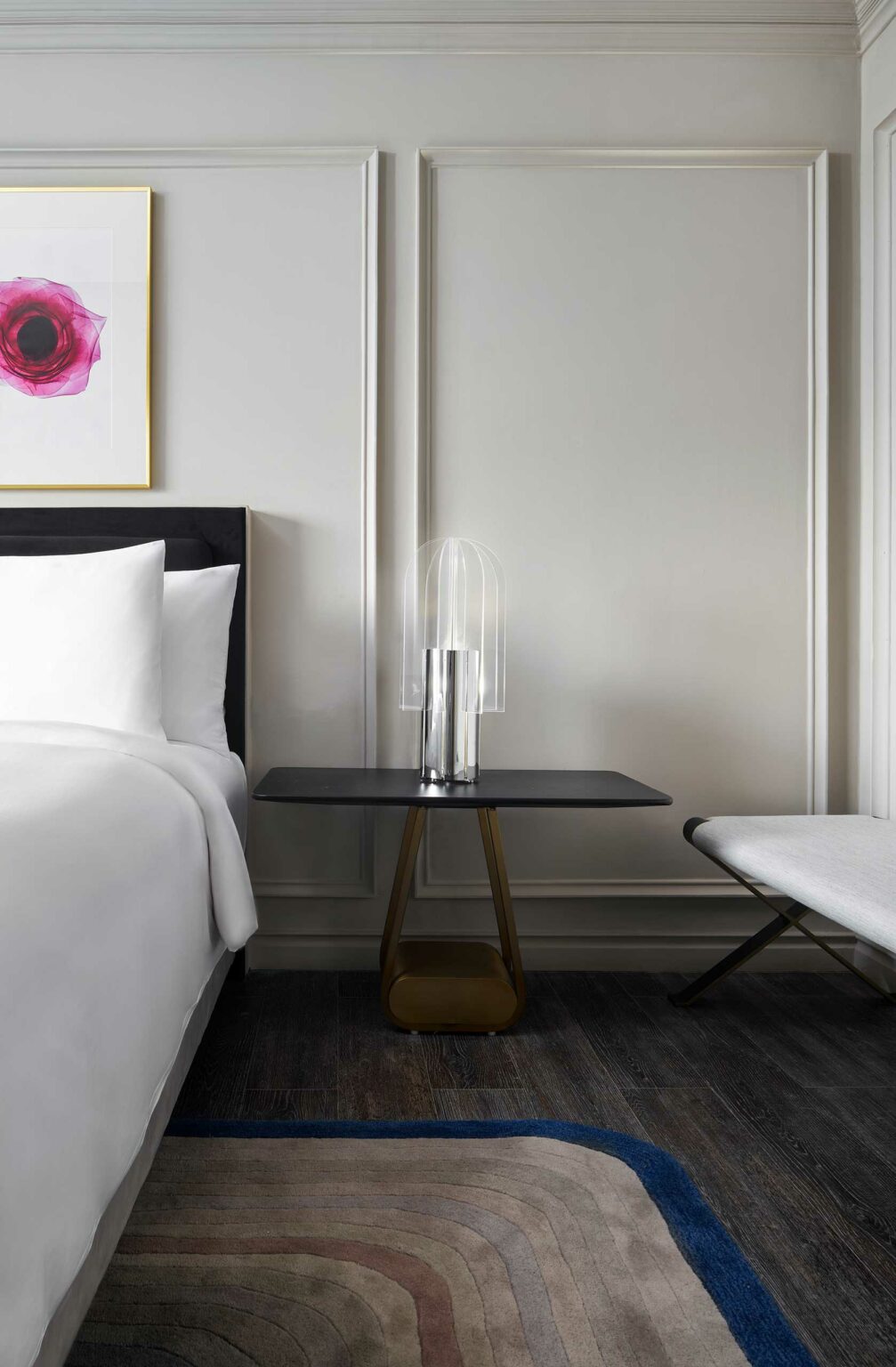 Stylish hotel bedroom with a modern vibe: a bed with white sheets, a sleek side table with a clear, curved lamp, abstract wall art, and a minimalist bench.
