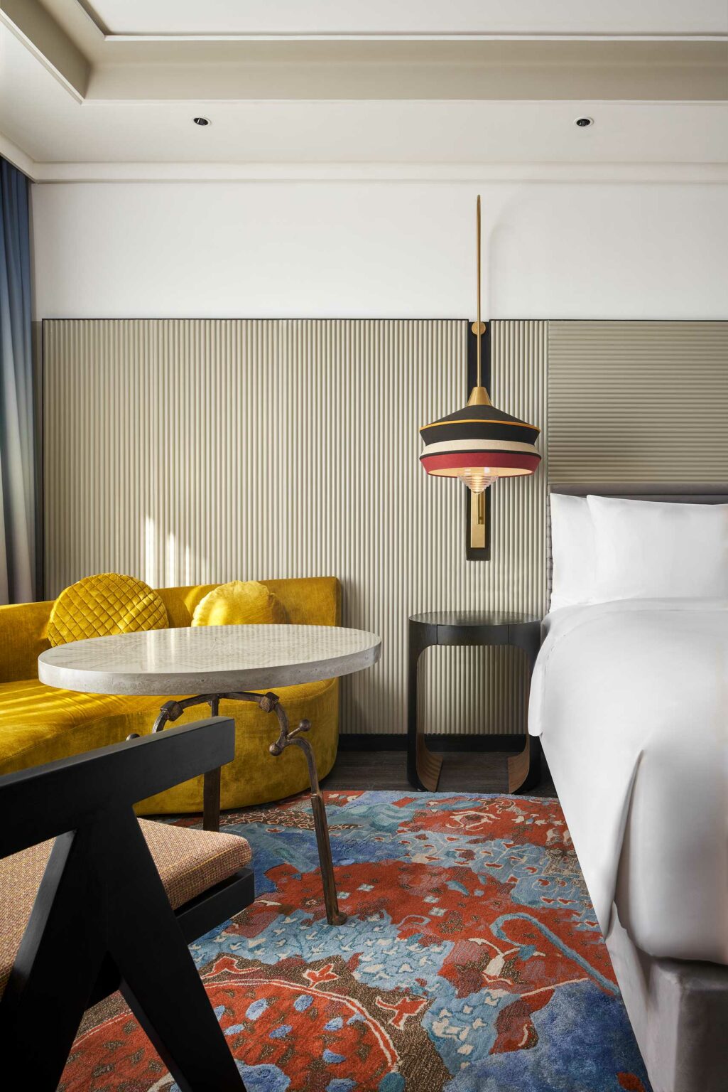 A modern hotel room features a white bed, a colorful pendant lamp, a wooden nightstand, a yellow chair, and a vibrant floral rug, creating a chic, cozy ambiance.