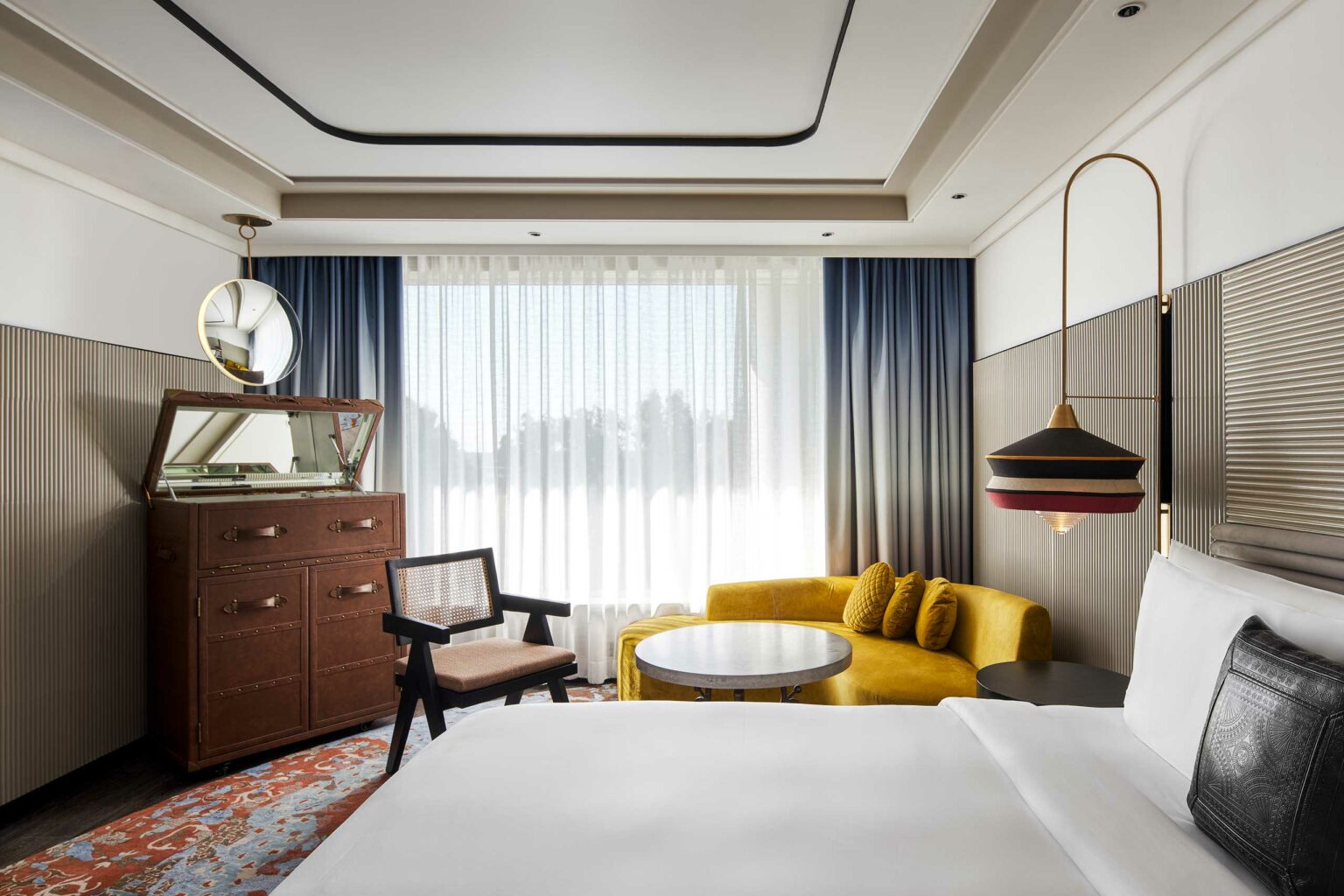 Modern hotel room with a cozy yellow sofa, round table, and vintage dresser. A large bed is in the foreground. Soft light filters through sheer curtains.