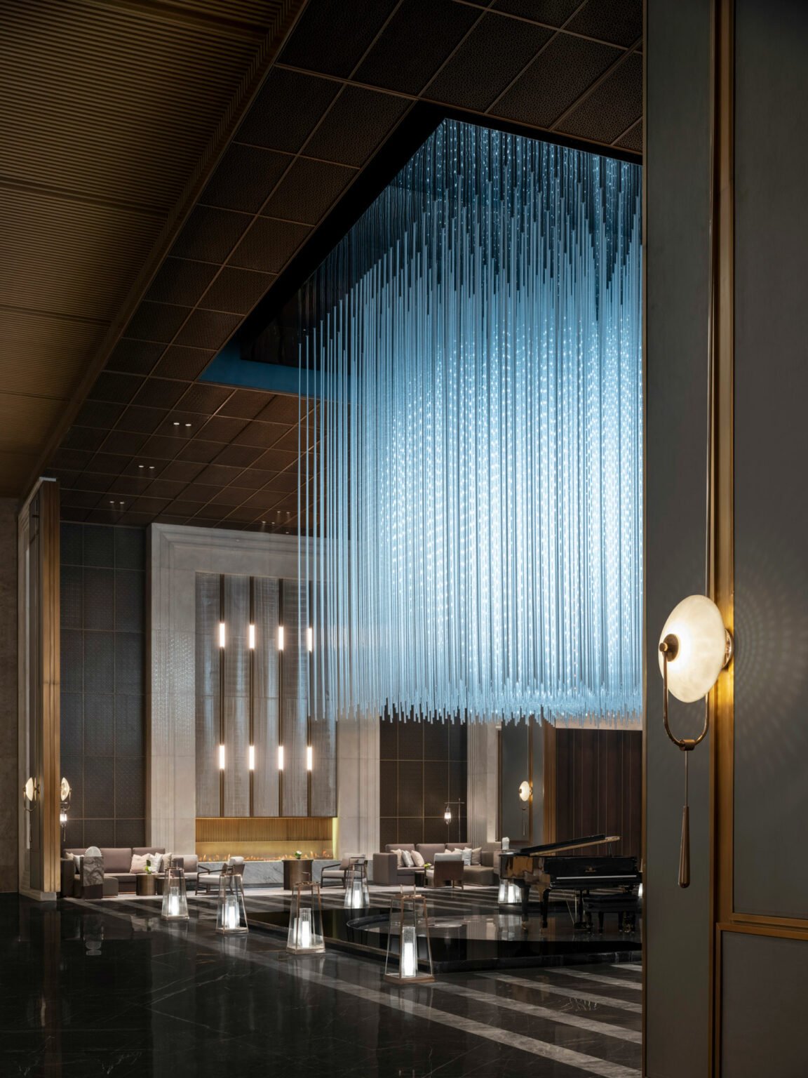 Luxurious hotel lobby featuring a grand, cascading blue chandelier, elegant seating, warm lighting, and a grand piano, creating a sophisticated ambiance.