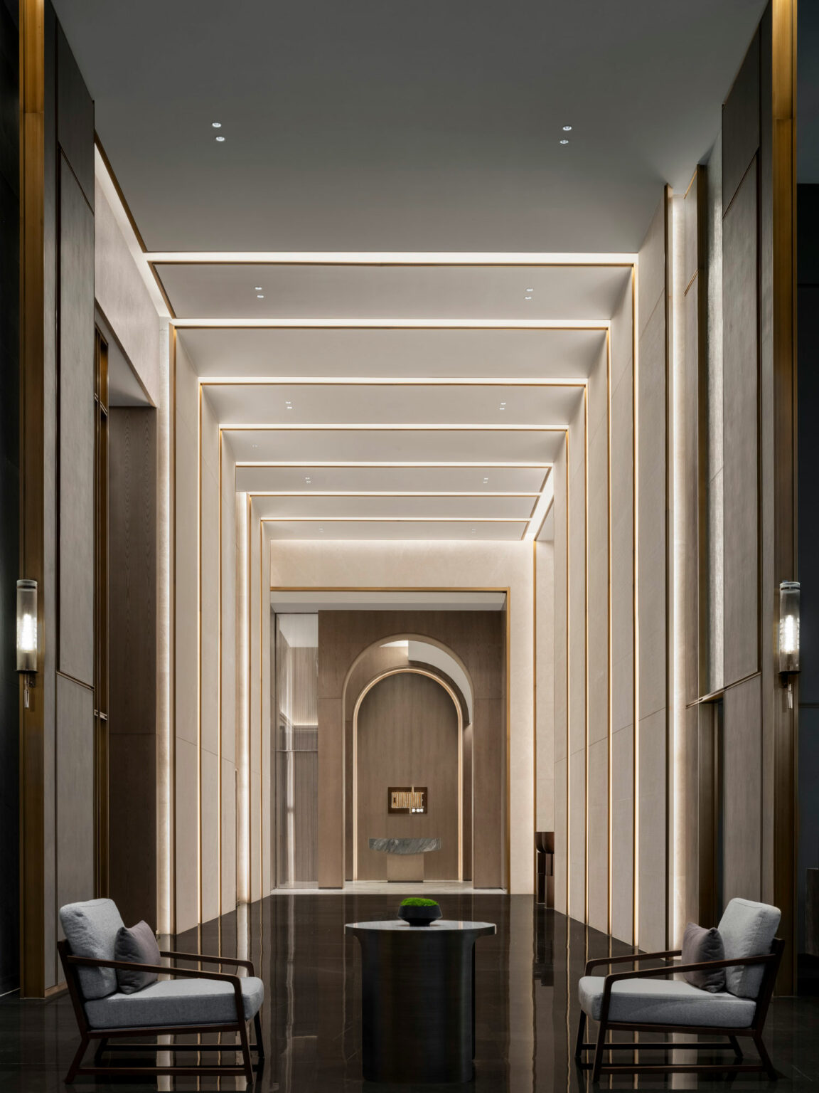 A modern, elegant hotel lift lobby with high ceilings, ambient lighting, and sleek wood paneling. Two armchairs face a round table in a serene setting.