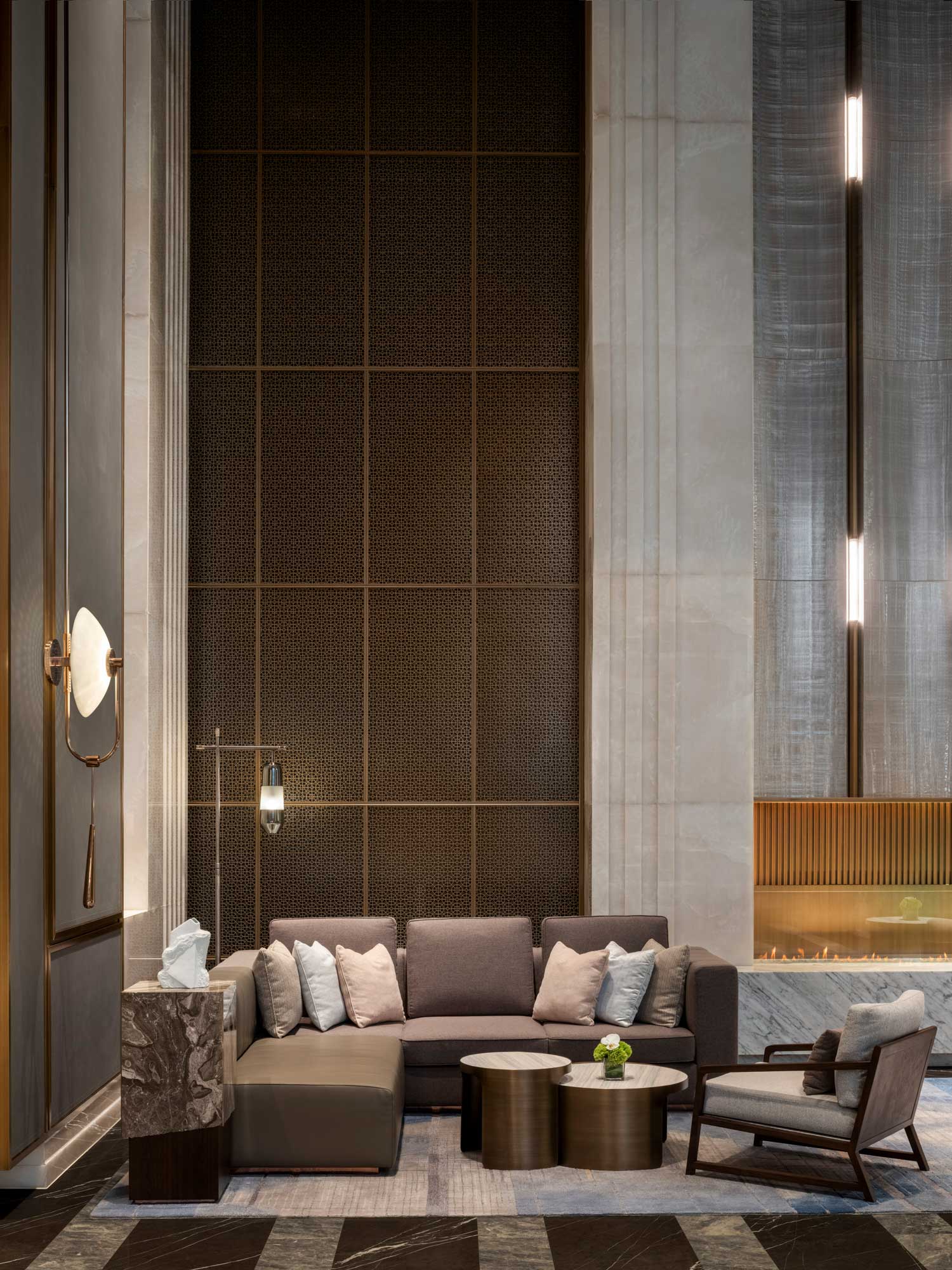 A modern, elegant hotel lounge with a sleek gray sectional sofa adorned with white pillows, a chic marble side table, and a minimalist fireplace.