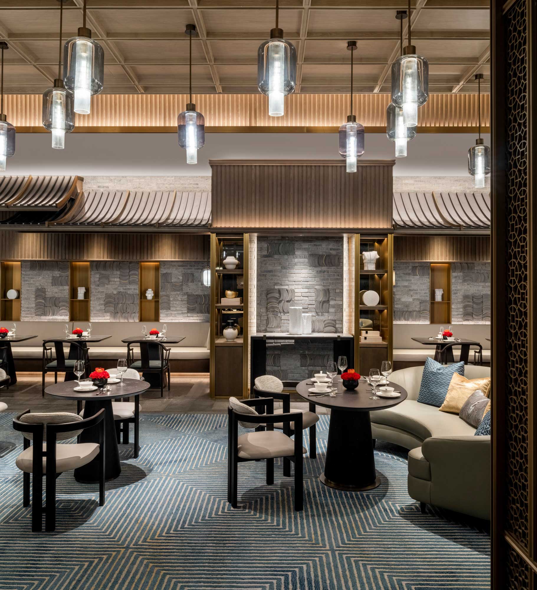 Elegant hotel all day dining with modern pendant lights, round tables set with glasses, and red floral décor. Cozy seating includes sofas and cushioned chairs on blue-patterned carpet. The ambiance is sophisticated and inviting.