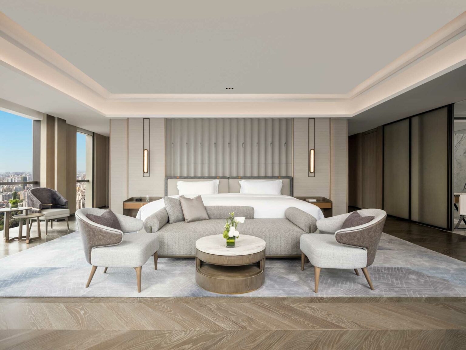 Spacious, modern hotel room with a large bed, plush gray seating, and round tables. Floor-to-ceiling windows offer a city view, creating a serene ambiance.