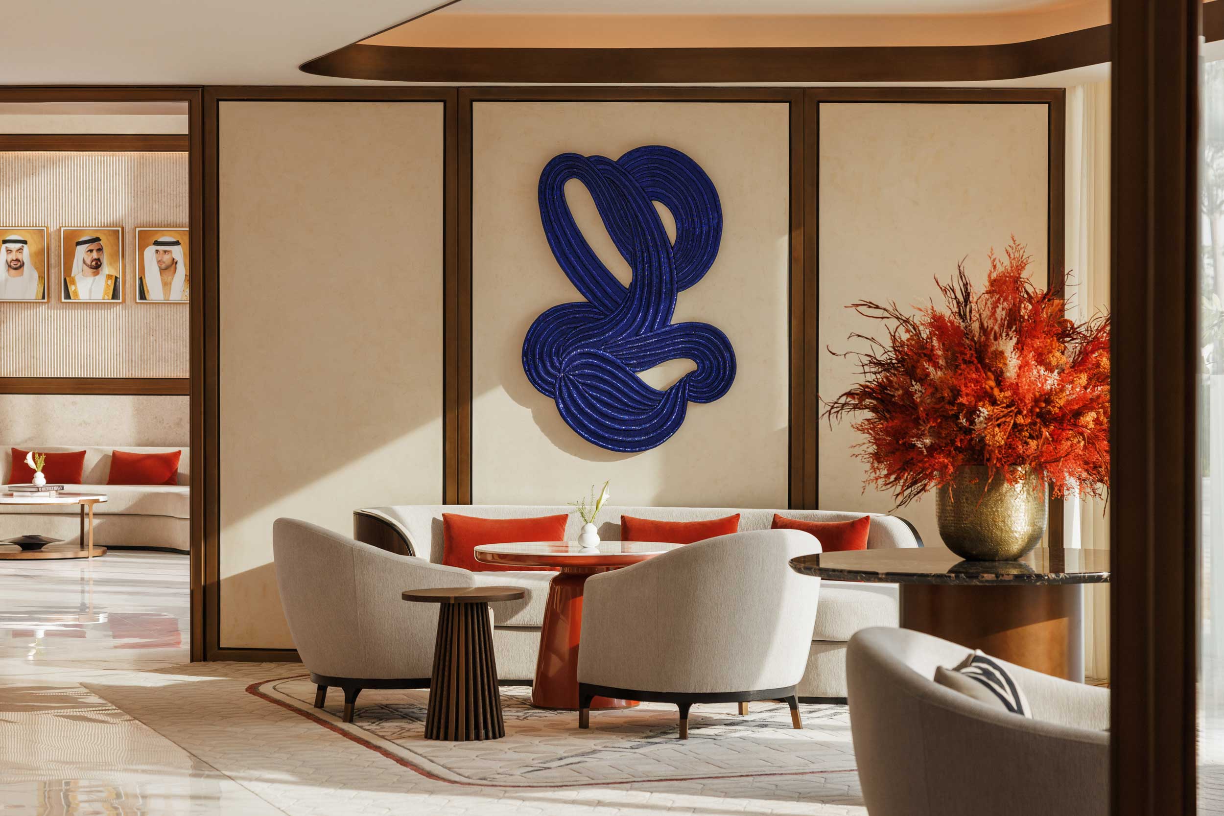 Modern hotel lounge with elegant neutral chairs, round table, vibrant red cushions, and a striking blue abstract wall art. Warm ambiance, decorative plant.