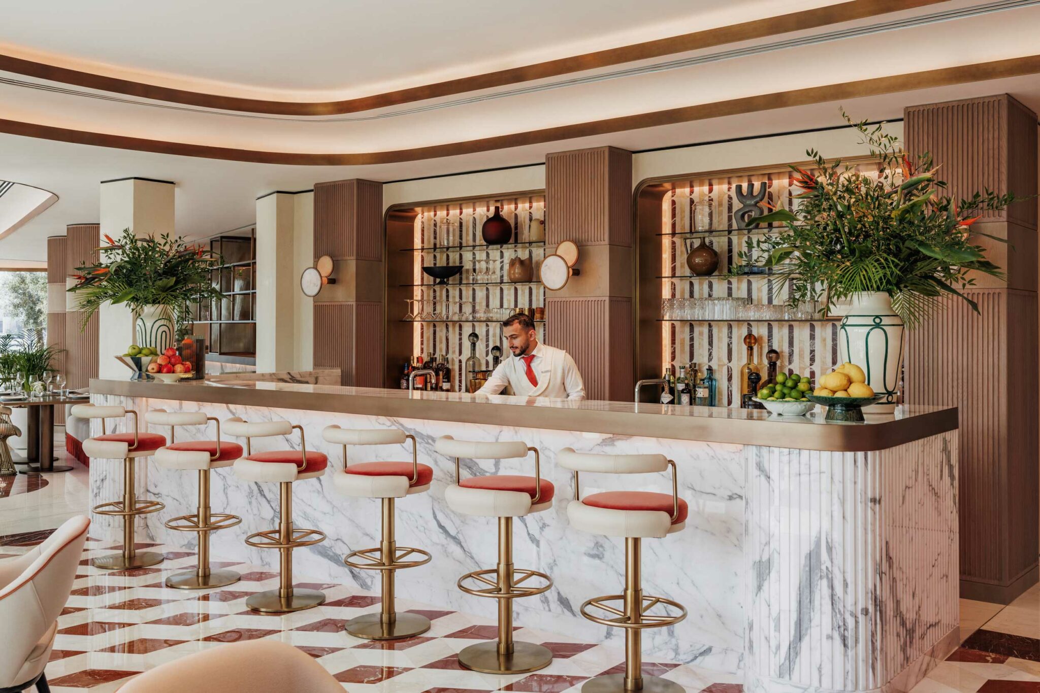 Elegant bar of a luxury hotel with a marble counter and red cushioned stools. A bartender prepares drinks amid lush greenery and fruit displays, creating a luxurious ambiance.