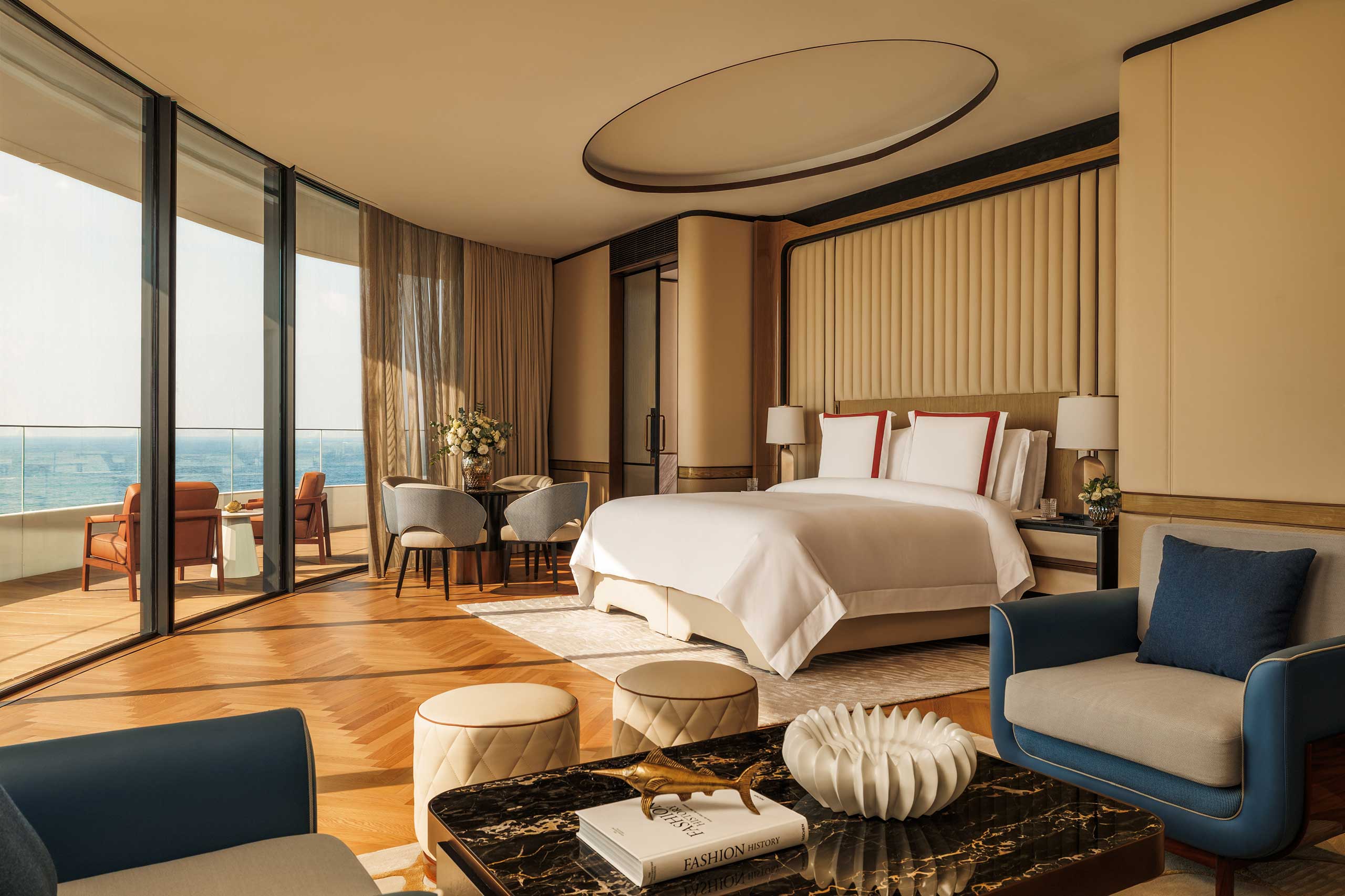 Luxurious hotel room with a king-sized bed, elegant decor, and large windows offering a stunning city view. Warm tones and plush seating create a cozy, sophisticated ambiance.