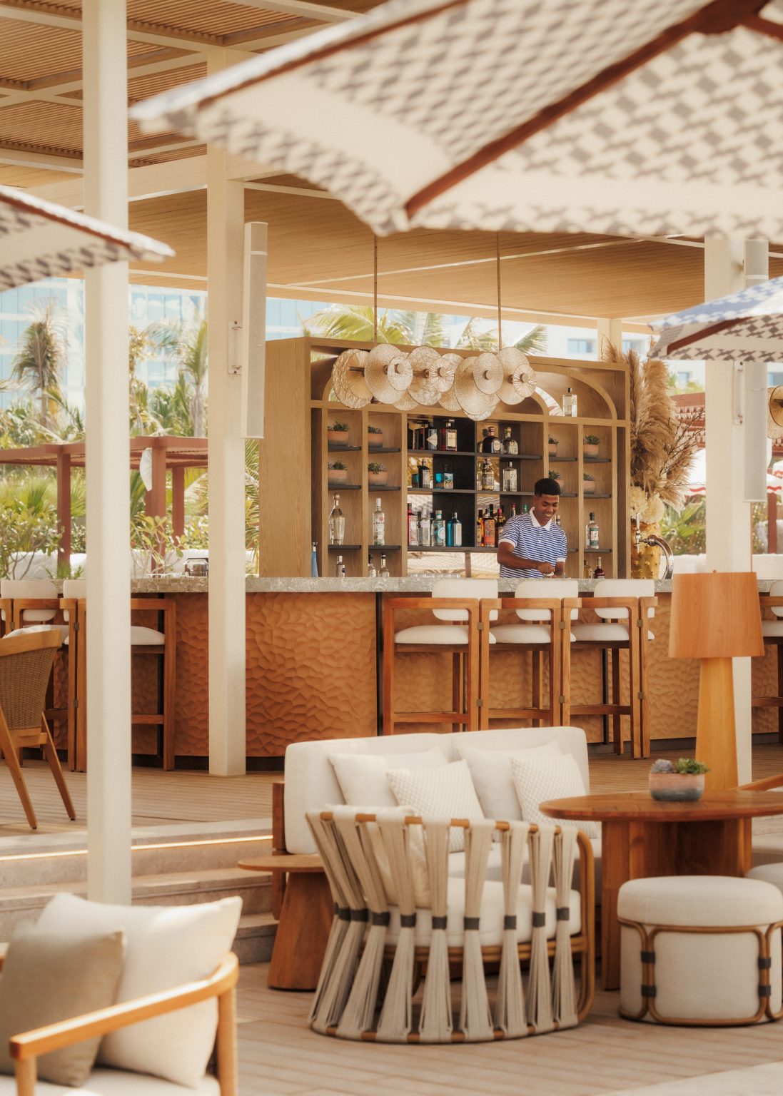 Chic outdoor bar with wooden stools, a variety of bottles on display, and stylish white umbrellas. The setting is bright and inviting, exuding a tropical vibe.