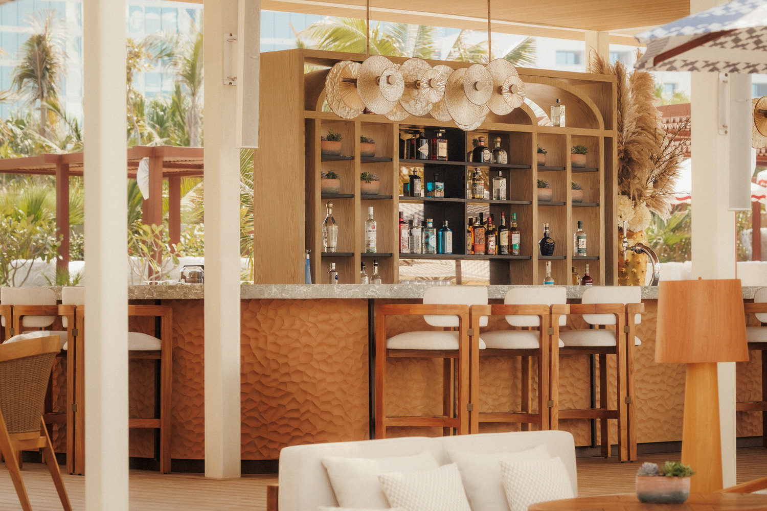 Chic outdoor bar with wooden stools, a variety of bottles on display, and stylish white umbrellas. The setting is bright and inviting, exuding a tropical vibe.
