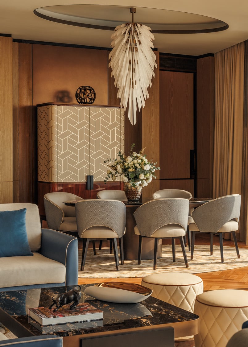 Elegant suite living room with a geometric-patterned cabinet, a round table surrounded by plush chairs, a central floral arrangement, and a modern chandelier.