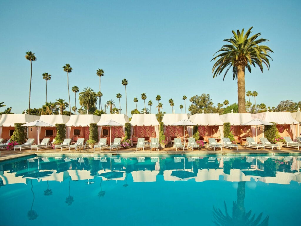 Poolside scene with striped lounge chairs and pink cabanas under umbrellas. Tall, lush palm trees line the background, evoking a serene, tropical vibe.