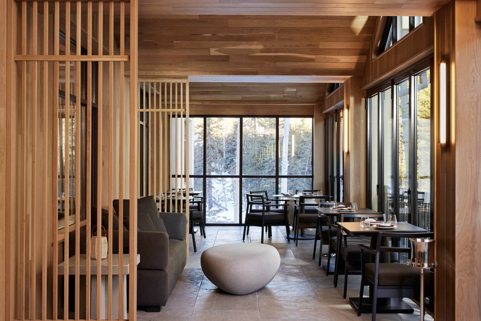 Modern restaurant interior with wooden paneling, large windows showing a snowy forest, gray seating, black tables, and soft ambient lighting. Cozy and inviting.