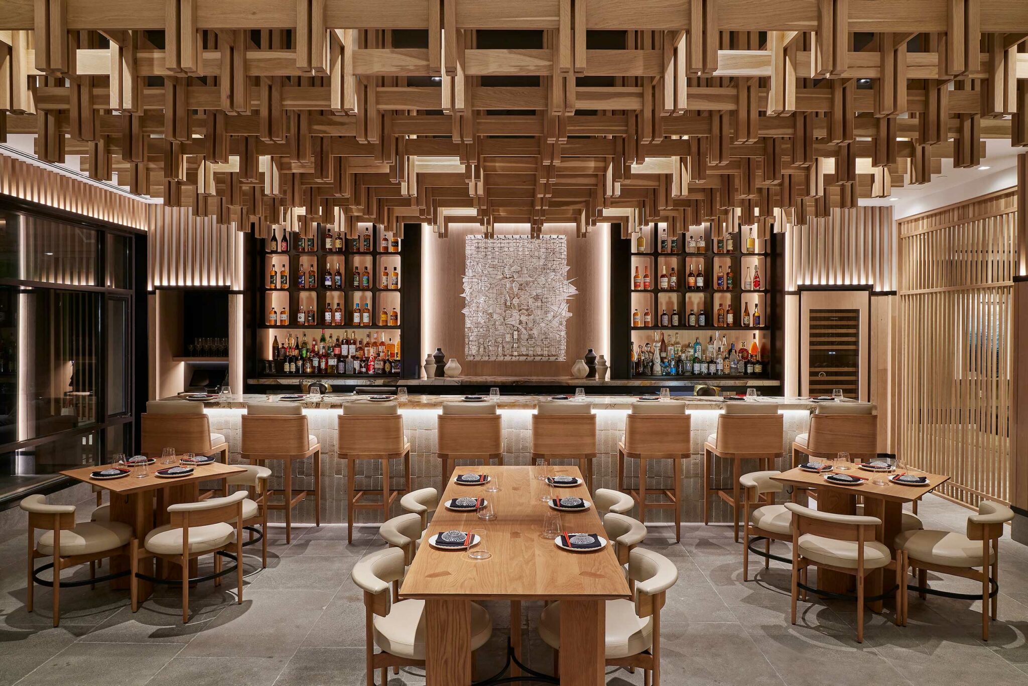 Warm, modern restaurant interior with wooden decor, geometric ceiling, and a central bar. Wooden tables and upholstered chairs offer an inviting atmosphere.