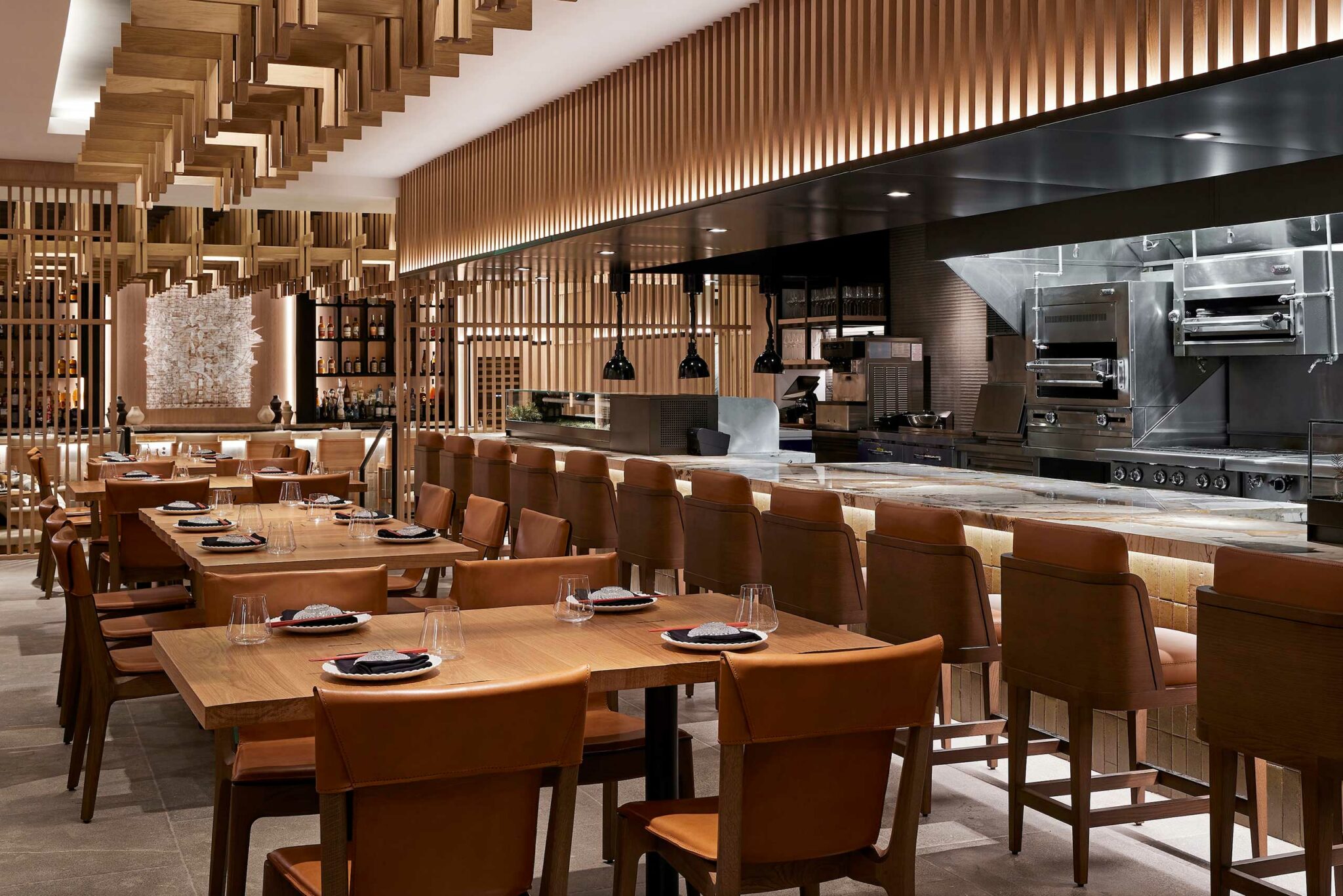 Modern restaurant interior with warm wood tones, leather chairs, and an open kitchen. The atmosphere is upscale and inviting, with neatly set tables.