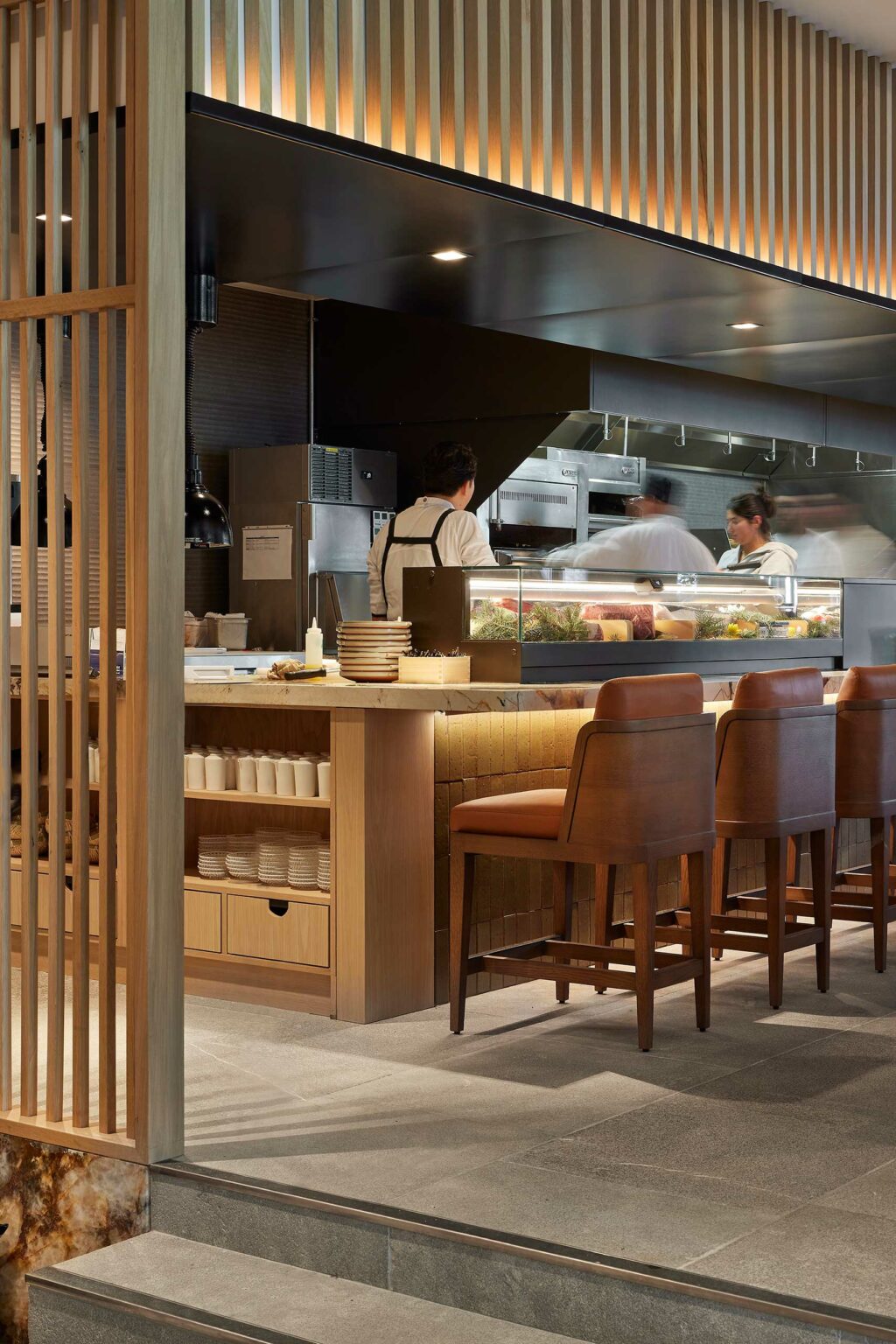 Open kitchen in a modern restaurant with chefs preparing food. Warm lighting, wooden accents, bar stools create an inviting and lively atmosphere.