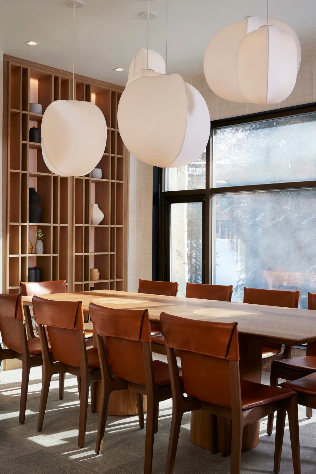 Modern restaurant dining room with a long wooden table, surrounded by leather chairs. Oversized white pendant lights hang above, and a wooden shelf with ceramics is in the background.