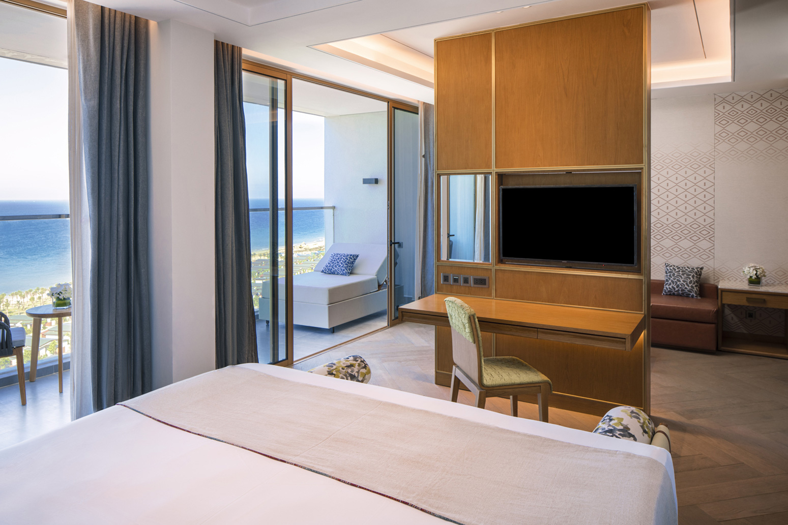 Modern hotel room with ocean view. Features a bed, wooden desk with a chair, wall-mounted TV, and a balcony with seating. Bright and relaxing ambiance.
