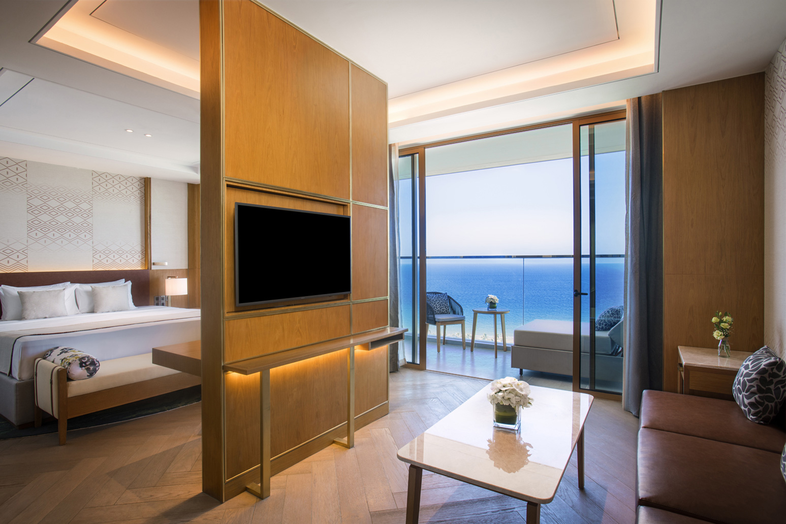 Modern hotel room with wooden decor, flat-screen TV, and cozy seating. A glass door reveals a serene ocean view, enhancing the tranquil ambiance.