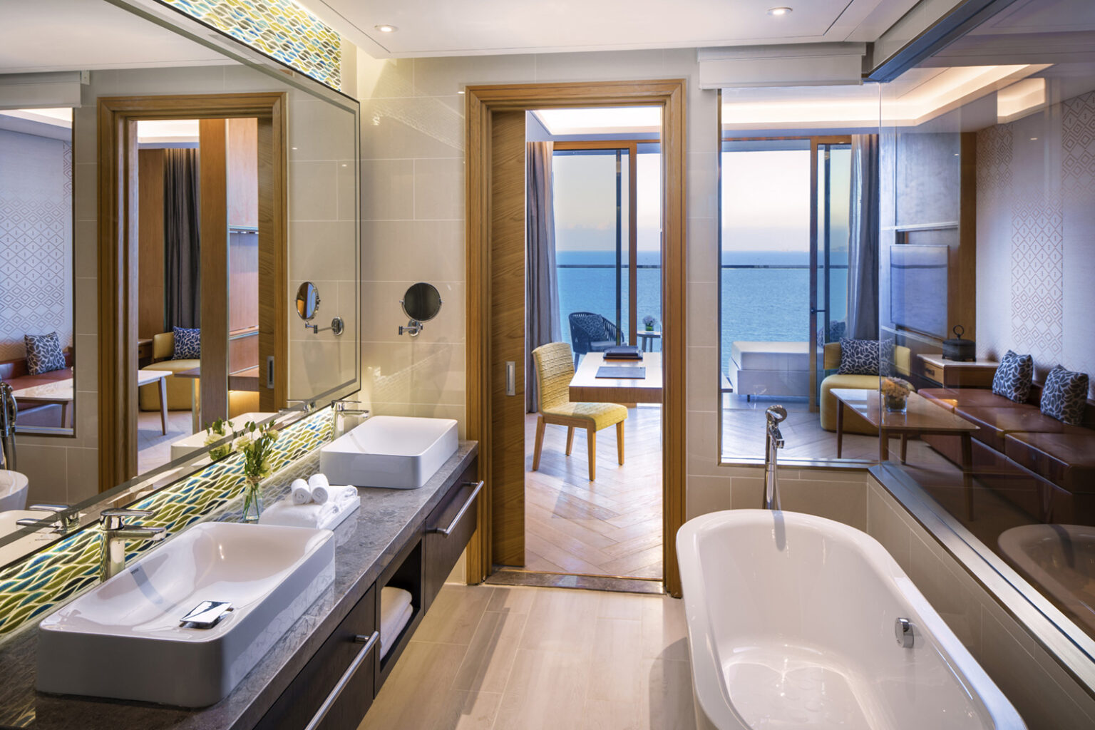 Modern bathroom with two sinks, a bathtub, and a view through a wooden door to a cozy room with ocean-view balcony. The scene is serene and luxurious.
