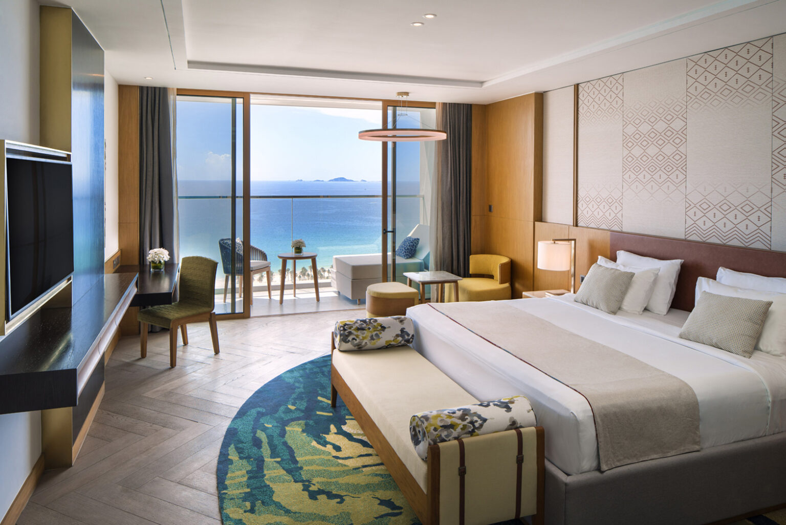 Hotel room with ocean view, king bed, and stylish decor. Floor-to-ceiling windows open to a balcony with seating. Calm and inviting ambiance.