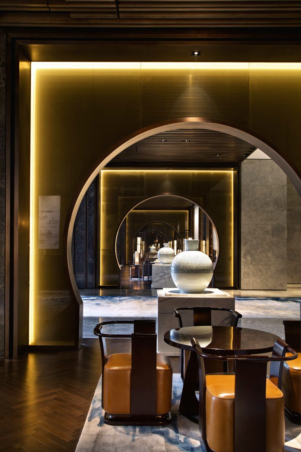 Luxurious, warmly lit hotel hallway featuring a series of archways with glowing trim. A round table and leather chairs stand in foreground, large vases display behind.