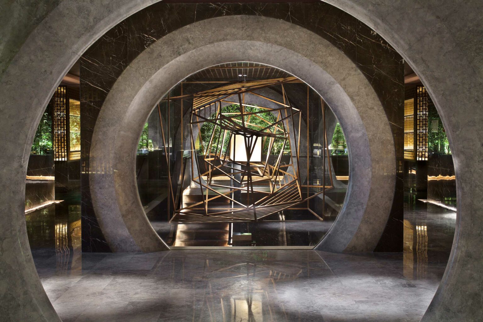 Circular stone entrance frames a geometric wooden sculpture in a modern hotel spa lobby. Sunlight filters through, highlighting green foliage outside.