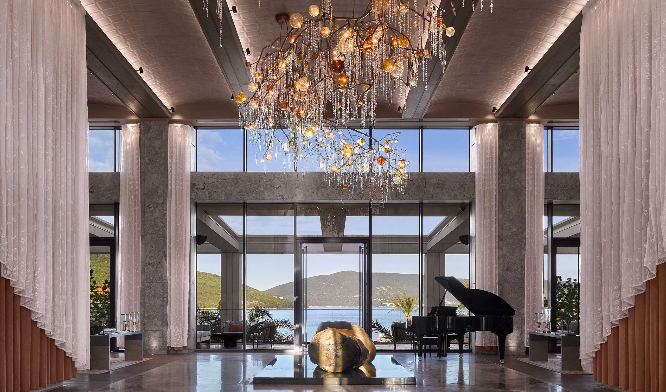 Spacious lobby with modern chandelier, large windows revealing a scenic view of hills and water. A grand piano adds elegance and sophistication.