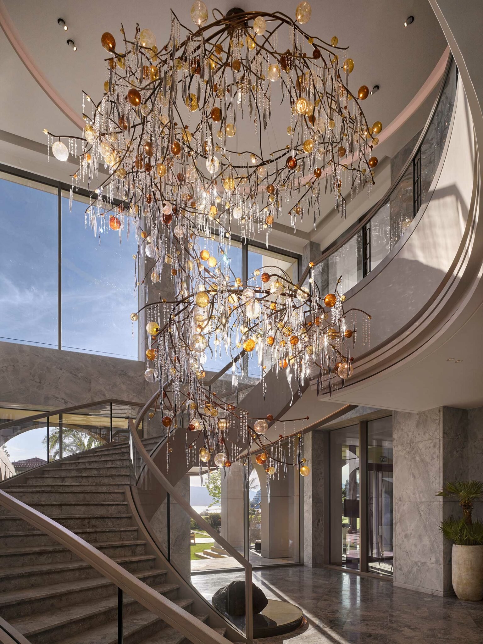 Elegant chandelier in a luxurious foyer with a grand spiral staircase. Glass orbs and crystals hang from branch-like metalwork, creating a radiant, opulent atmosphere.