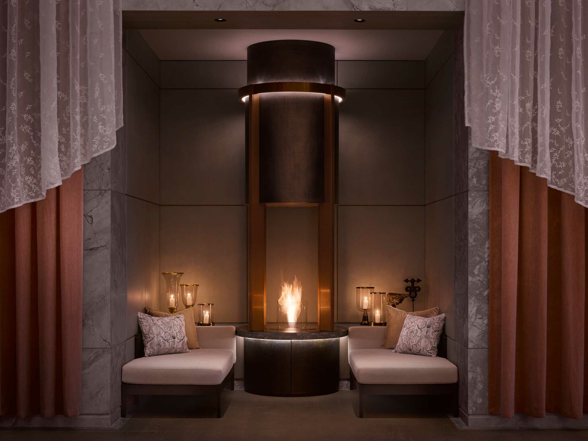Luxurious, intimate resort lounge with a central flame feature flanked by two cushioned benches. Soft lighting and ornate curtains create a warm, serene atmosphere.