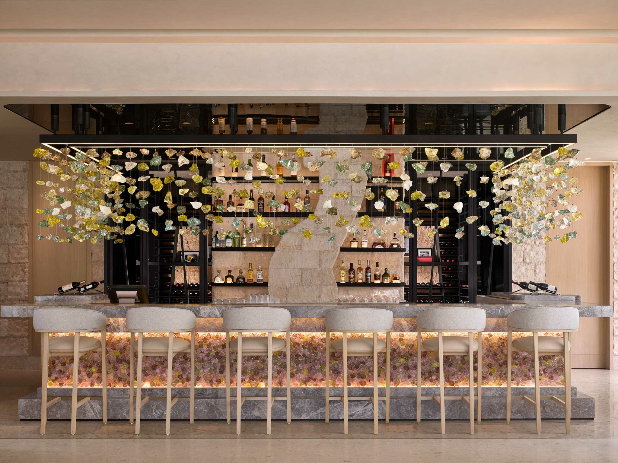 Elegant resort bar with a modern stone counter, plush bar stools, and a backlit floral design. Glass decorations hang above, with shelves of neatly arranged bottles.