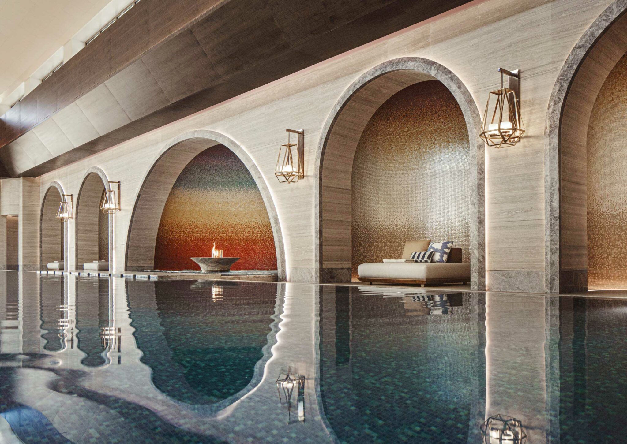 Luxurious indoor pool with reflective water, flanked by elegant stone arches. A cozy seating area and a lit fire bowl create a serene, upscale ambiance.
