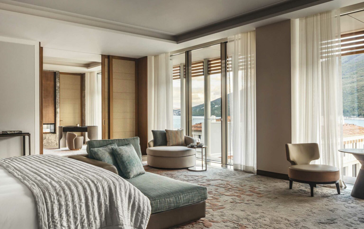 Luxurious resort room with modern decor, featuring a plush bed, lounge chairs, large windows with sheer curtains, and a scenic view of mountains and water.