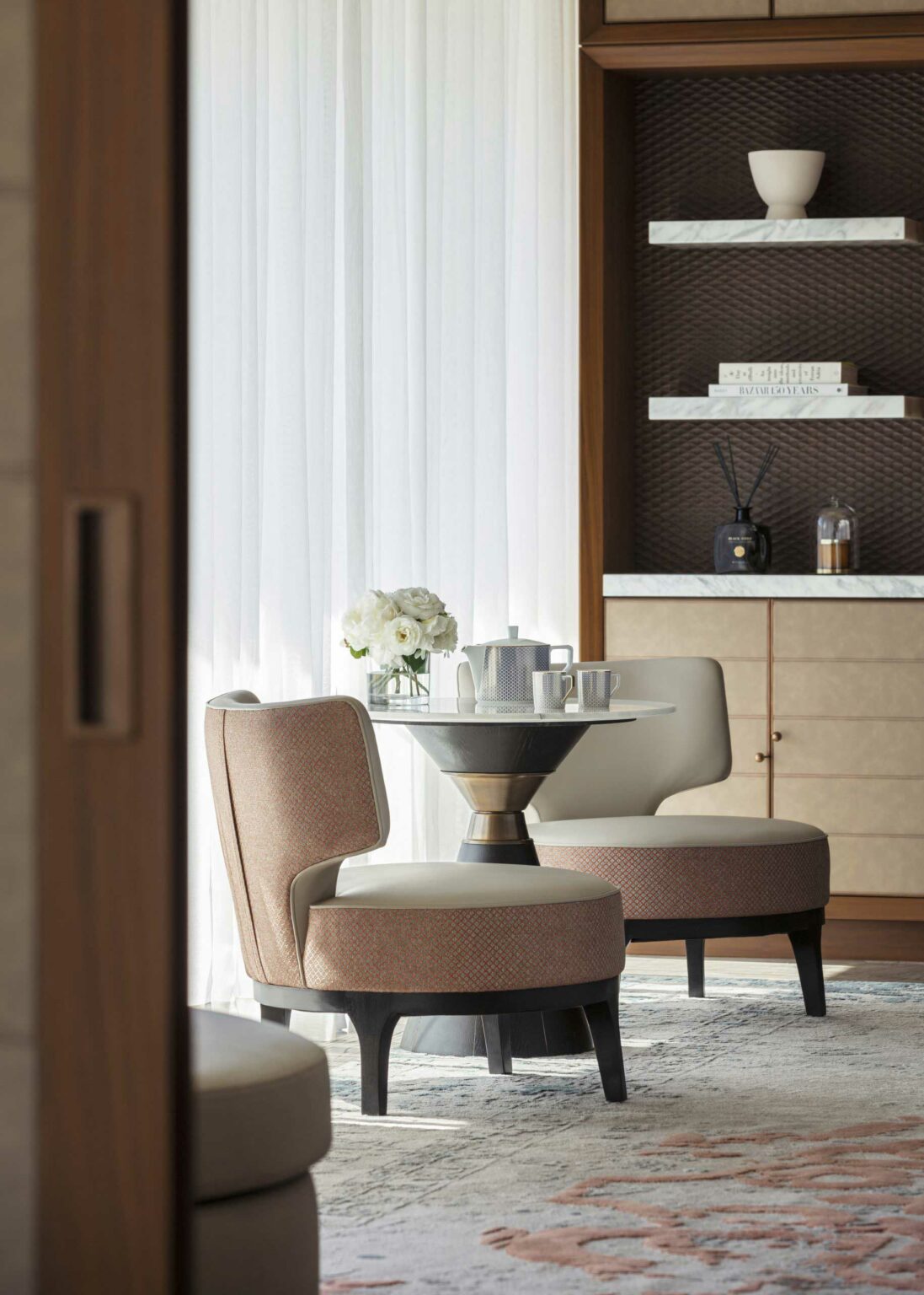 Cozy sitting area with two cushioned chairs and a round table set with a teapot and cups. White curtains softly filter light. Warm, elegant ambiance.