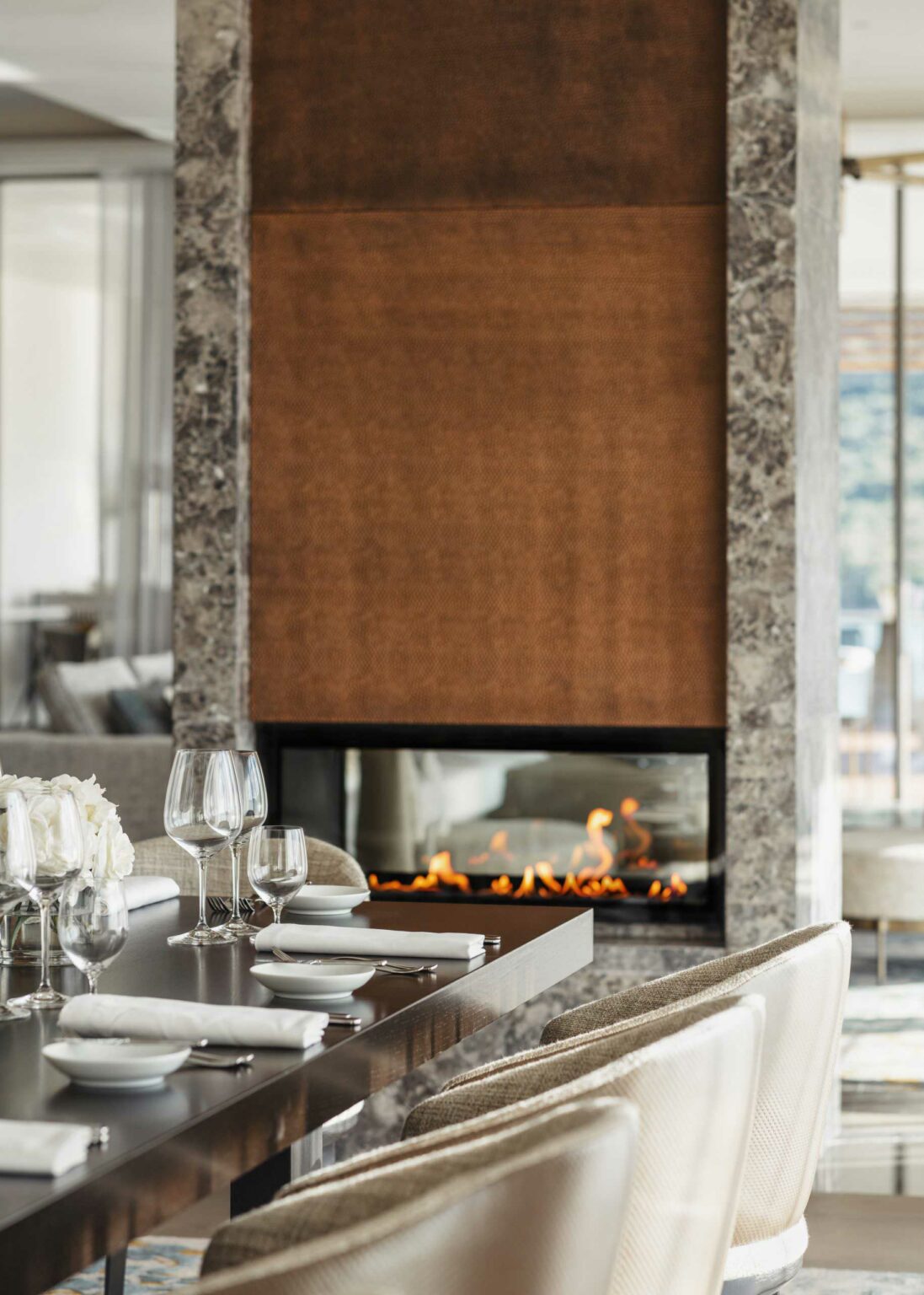 Elegant resort dining area with a modern fireplace, tall flames flickering behind glass. Table set with white linens, glasses, and plush beige chairs, evoking warmth.
