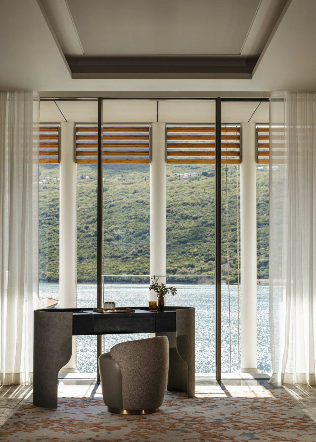 Modern suite study room with floor-to-ceiling windows showing a scenic view of a lake and green hills. A stylish desk and chair are in the foreground under soft light.