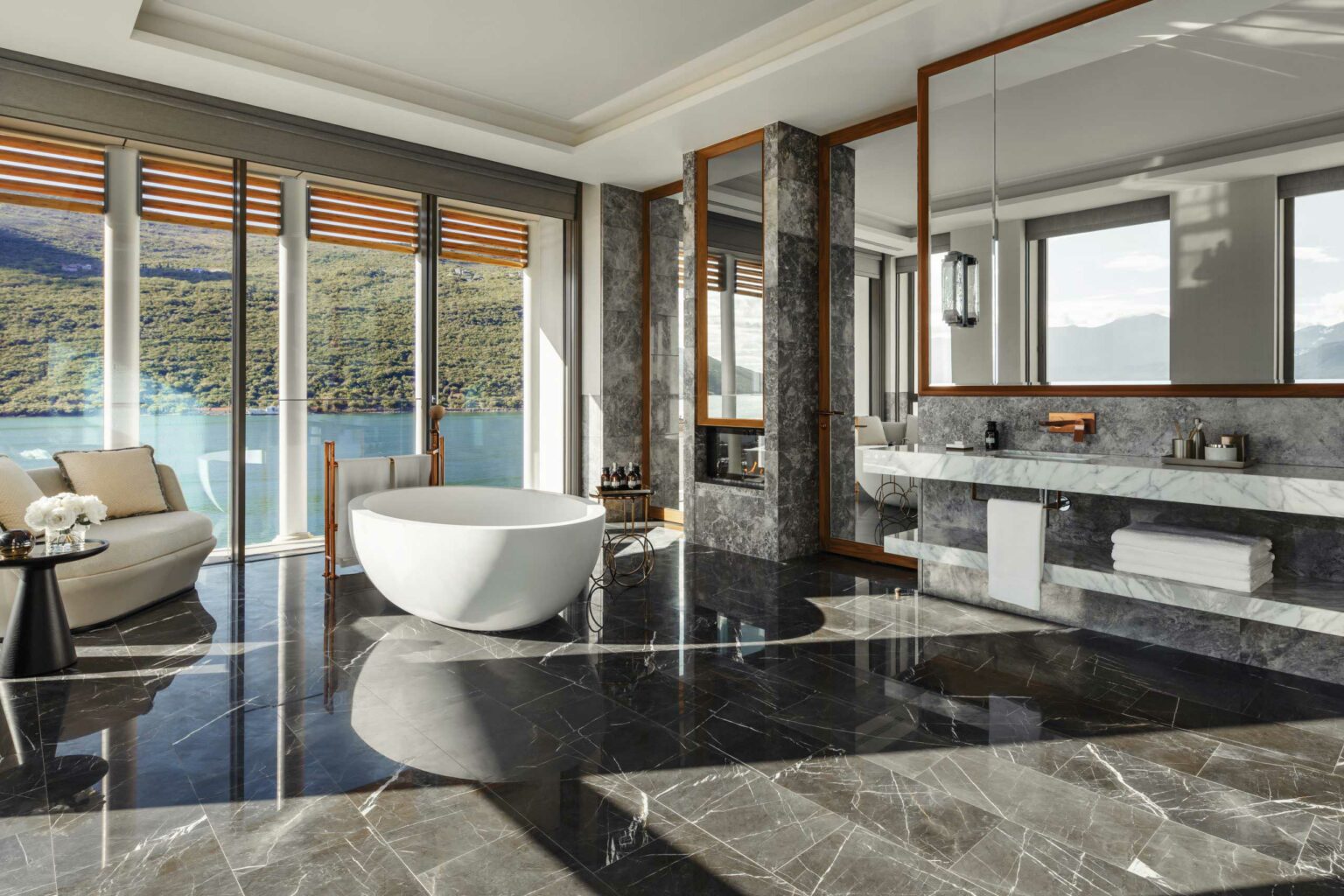 Luxurious suite bathroom with a freestanding tub, marble floors, and large windows offering a scenic view of a lake and hills, creating a serene ambiance.