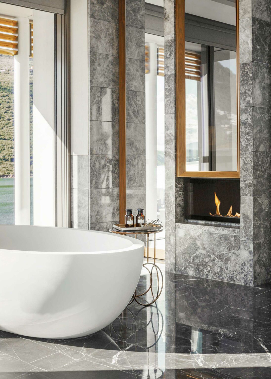 Luxurious resort bathroom with a white oval bathtub, marble floors, and walls. A small round table holds toiletries, next to a modern fireplace. Bright and serene.