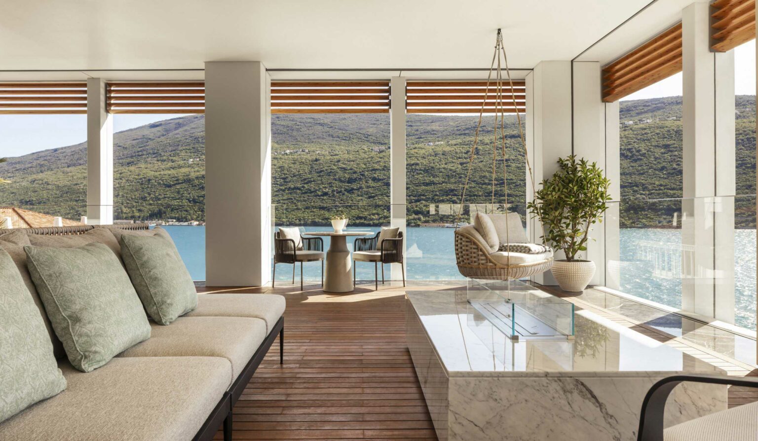 Modern suite living room with large windows showing a lake and mountains. Features a swing chair, wooden floor, potted plant, and elegant furniture. Relaxing atmosphere.