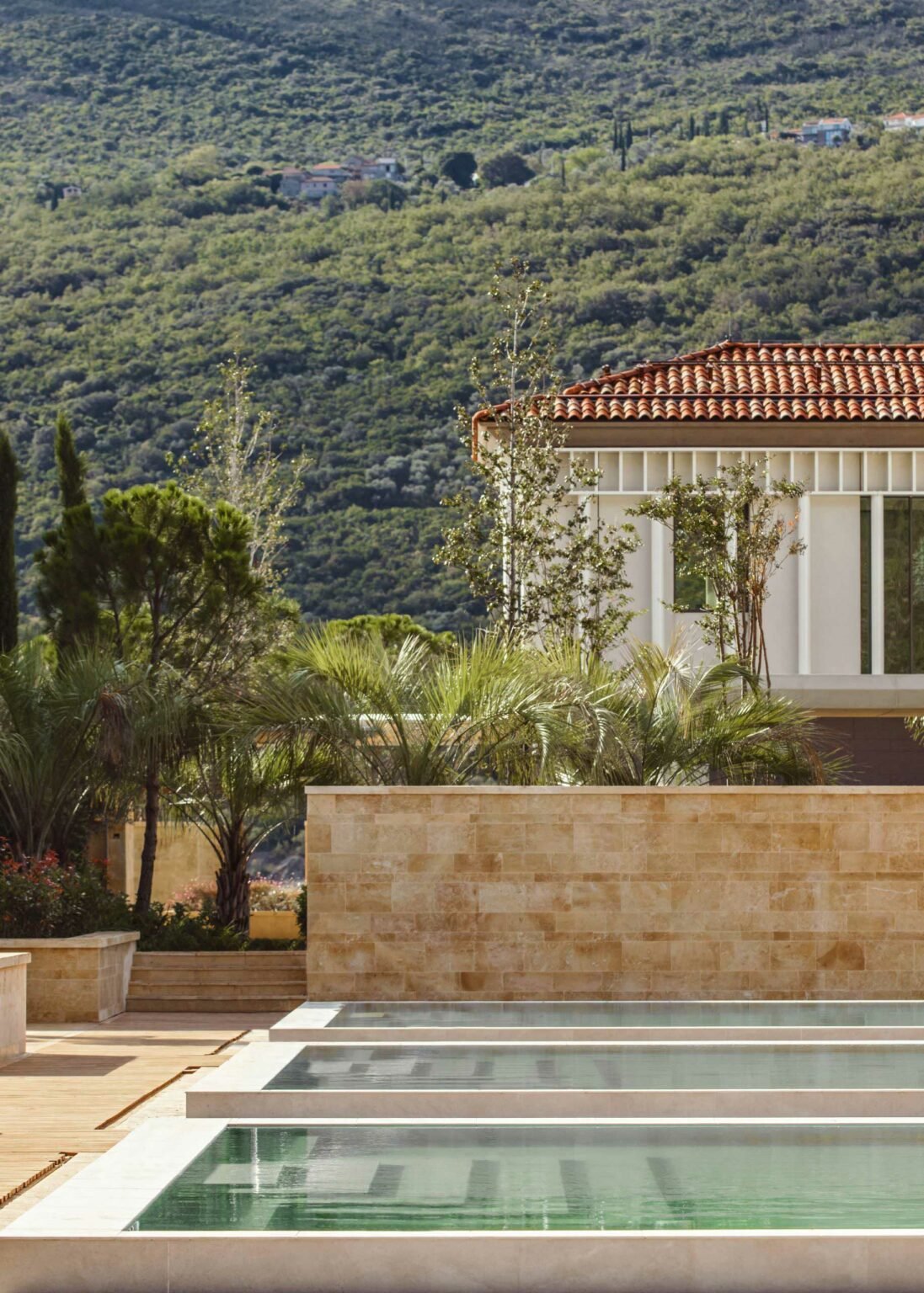 Luxurious villa with terracotta roof, palm trees, and a serene infinity pool in the foreground. Backdrop of lush green hills, evoking tranquility.