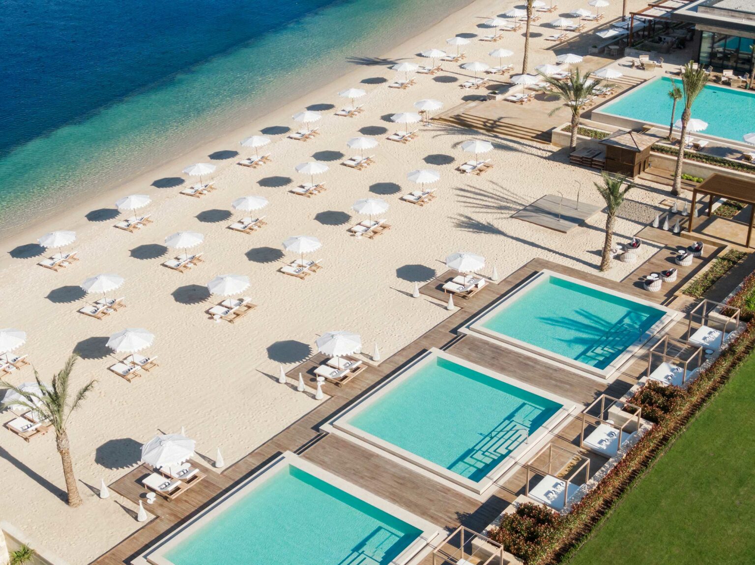 Aerial view of a serene beach with rows of white umbrellas and loungers on golden sand, adjacent to three turquoise pools, creating a tranquil resort ambiance.